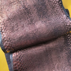 Puede incluir: Un trozo de cuero de color bronce metálico con un patrón texturizado y en relieve de piel de serpiente. El material tiene un acabado brillante y reflectante con tonos marrón oscuro visibles en las escamas, mostrando un borde doblado.