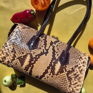 May include: A rectangular handbag featuring a textured snakeskin pattern in shades of tan, beige, and dark brown. The bag includes two dark brown, braided-style shoulder straps attached with triangular patches and a gold-toned zipper closure.