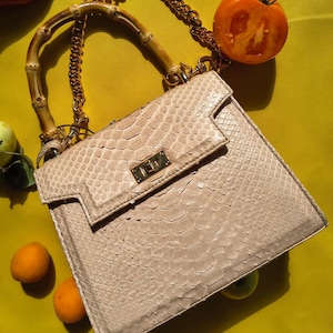 May include: A beige snakeskin-textured handbag featuring a bamboo top handle and a gold-tone metal chain shoulder strap. The bag has a structured rectangular shape with a front flap closure secured by a gold-tone rectangular twist lock clasp.