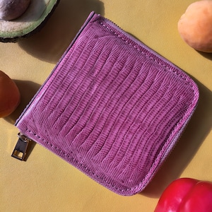 Peut inclure: Un portefeuille carré de couleur violette présentant une texture en relief façon peau de lézard. Le portefeuille est doté d'une fermeture à glissière sur deux côtés, terminée par une tirette métallique de couleur argentée sur le coin.