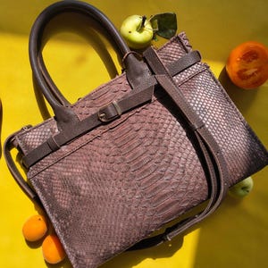 Peut inclure: Un sac à main marron avec un motif texturé en peau de serpent, doté d'une poignée en cuir lisse, d'une fine bandoulière réglable et d'une ceinture en cuir horizontale sur le devant, fixée par une petite boucle en métal et un passant.