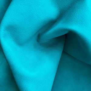 Puede incluir: Una piel de cuero de color turquesa vibrante con una textura de grano natural distintiva. El material está doblado para crear sombras y reflejos profundos, mostrando la superficie lisa y mate y el color azul verdoso brillante y uniforme.