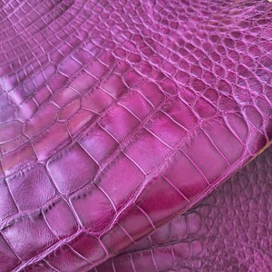 May include: A vibrant magenta purple leather hide featuring a distinct embossed crocodile skin texture. The material displays a glossy finish with deep, realistic scale patterns that vary in size across the surface, creating a dimensional look.