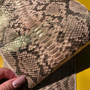 May include: A piece of textured material featuring a metallic gold and black snakeskin pattern. The surface displays a repeating scale design with a reflective sheen, showing a detailed reptilian texture across the entire visible surface area.