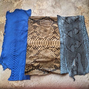 May include: Three rectangular pieces of textured leather with a snake scale pattern. The left piece is vibrant blue, the center piece is tan and brown with a natural scale design, and the right piece is a muted grey-blue with dark markings.
