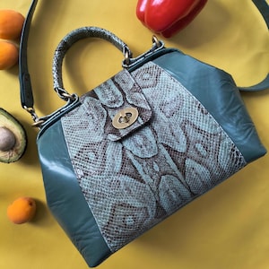 May include: A structured handbag featuring a teal leather body and a central panel with a textured snakeskin pattern in shades of brown and light blue. The bag includes a top handle, a detachable shoulder strap, and a gold-tone metal twist lock.