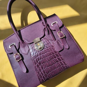 Puede incluir: Un bolso de mano de color morado oscuro con acabado texturizado y un panel central vertical con patrón de cocodrilo en relieve. El bolso incluye dos asas superiores, un cierre central de metal y dos correas laterales con hebillas doradas.