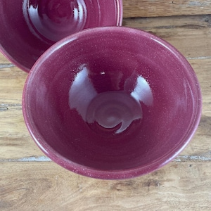May include: Two deep, round bowls in a speckled, glossy burgundy glaze. The bowls are nested, with one partially visible behind the other. The interior of the bowls reflects the light, highlighting their smooth surfaces. The bowls are likely for serving food.