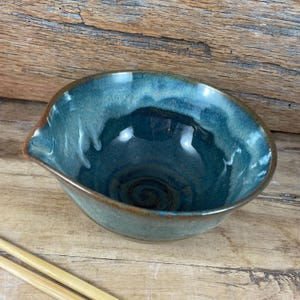 Può includere: Una ciotola in ceramica fatta a mano con smalto turchese e bordo marrone. La ciotola ha un beccuccio ed è appoggiata su una superficie di legno. La ciotola è vuota.