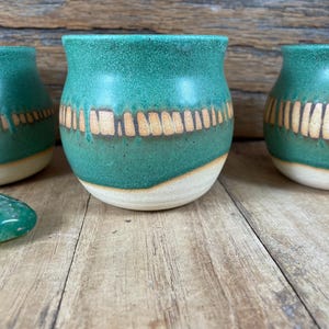 May include: Three handmade ceramic cups with a teal glaze and a band of vertical tan lines. The cups have a rounded shape and a cream-colored base. The cups are displayed on a wooden surface.