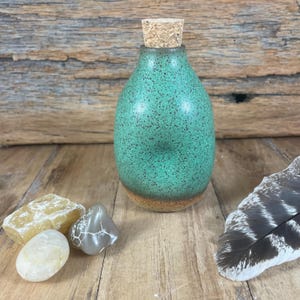 May include: A speckled teal ceramic bottle with a cork stopper. The bottle is displayed with several stones and a feather on a wooden surface. The bottle has a rounded shape and a speckled texture.