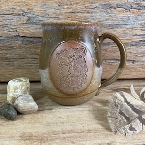 May include: A brown ceramic mug with a handle. The mug has a brown and white glaze and a raised design of a woman holding a baby on the front. The design is surrounded by a brown oval.