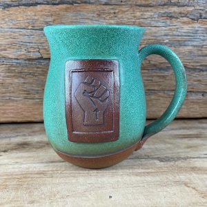 May include: A handmade ceramic mug with a green speckled glaze. A brown rectangular emblem with a raised fist design is centered on the mug. The handle and base are a darker shade of green and brown.