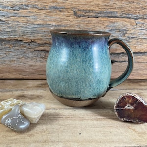 May include: A handmade ceramic mug with a rounded body and a large handle. The mug features a gradient of blue and green hues, with a brown rim and base. The mug is displayed on a wooden surface with decorative stones.