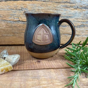 May include: A dark blue ceramic mug with a brown glaze and a raised triangle design. The triangle has a stylized symbol inside.