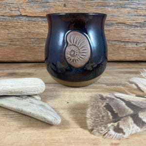 May include: A rounded ceramic mug with a glossy dark blue and black glaze, featuring a raised, unglazed tan clay emblem of a spiral ammonite fossil on the front. The base of the mug is exposed, unglazed tan clay, contrasting with the dark body.