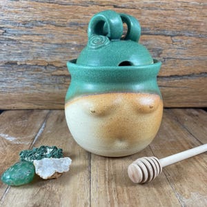 May include: A handmade ceramic honey pot with a green lid and a tan and cream-colored body. The lid has a handle with a spiral design. A wooden honey dipper rests beside the pot. Decorative stones are in the foreground.