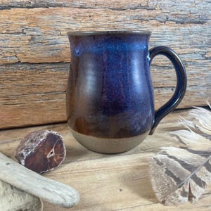 May include: A ceramic mug with a rounded body and a curved handle. The glaze features a deep blue and purple gradient at the top that transitions into a warm brown tone, ending in a light beige, unglazed base with a speckled texture.
