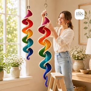 May include: Two spiral-shaped hanging decorations made of crocheted yarn in a vibrant rainbow gradient, featuring a cream-colored border. Each piece is suspended from a cord and a metal hook, displaying a continuous color transition from deep purple to red.