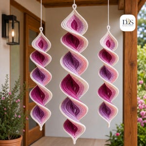Könnte beinhalten: Drei hängende Häkel-Spiralornamente mit einem Farbverlauf, der von tiefem Magenta und Violett zu hellem Rosa und Creme übergeht. Jedes Stück ist an einer weißen Schnur befestigt und zeigt ein strukturiertes, gedrehtes Design.