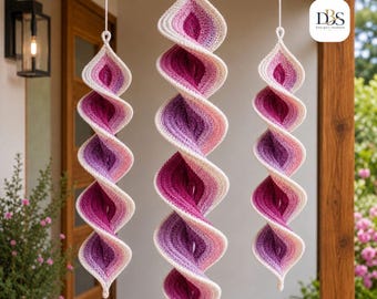 Op de afbeelding: Drie hangende gehaakte spiraalornamenten met een kleurverloop dat varieert van diep magenta en paars tot lichtroze en crème. Elk stuk hangt aan een wit koord en heeft een getextureerd, gedraaid ontwerp.