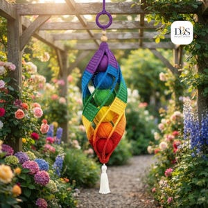 May include: A hanging crochet yarn holder featuring a spiral design in a rainbow color gradient. The structure holds five round balls of yarn in purple, blue, green, yellow, and red, suspended by a purple ring and finished with a white tassel.