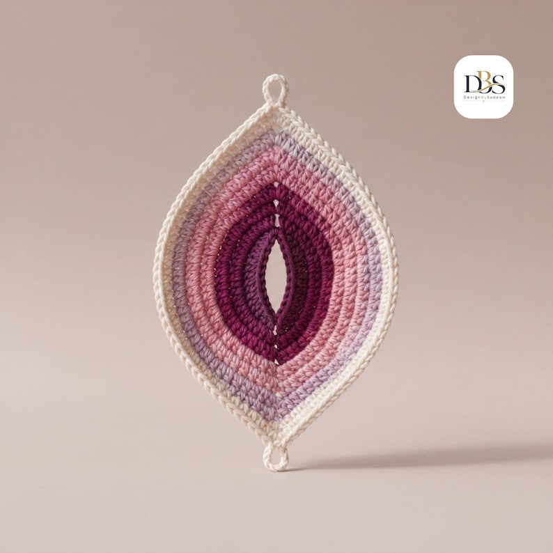 May include: A leaf-shaped crochet ornament featuring concentric layers of yarn in shades of cream, light purple, pink, and deep magenta. The piece has a small loop at the top and bottom for hanging and includes a small logo reading DBS Design.