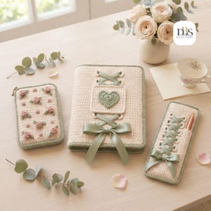 May include: A cream-colored crochet notebook cover with a sage green ribbon lace-up design and a central heart motif, accompanied by a matching crochet pouch with pink floral embroidery and a slim crochet pen case with a sage green ribbon bow.
