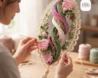 Puede incluir: Un tapiz decorativo de ganchillo con un marco ovalado de encaje color crema y una espiral central de tela verde, rosa y beige. La pieza está adornada con flores de ganchillo rosas y hojas verdes, rematada con una cuenta de madera y una borla.