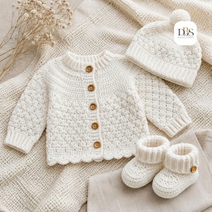 Peut inclure: Un ensemble en crochet couleur crème composé d'un gilet à manches longues avec cinq boutons marron, d'un bonnet assorti avec un pompon et d'une paire de chaussons avec un bouton marron sur chacun. Le texte DBS Design-Season est visible.