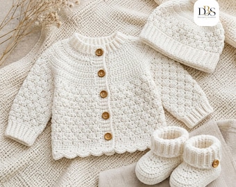 Puede incluir: Un conjunto de ganchillo color crema que consta de un cárdigan de manga larga con cinco botones marrones, un gorro a juego con un pompón y un par de patucos con un botón marrón en cada uno. El texto DBS Design-Season es visible.