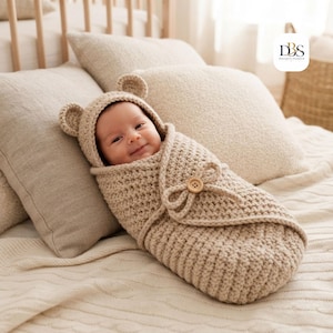 Peut inclure: Une couverture d'emmaillotage pour bébé en crochet beige dotée d'une capuche avec deux petites oreilles d'ours arrondies. L'emmaillotage enveloppe un nourrisson et est fixé par un bouton en bois brun clair à l'avant, offrant un aspect texturé.