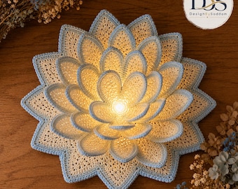 Puede incluir: Un aplique de pared con forma de flor de loto tejida a ganchillo, con pétalos superpuestos de color crema y bordes azul claro. El centro de la flor brilla con una luz cálida. Un logotipo circular en la esquina superior derecha muestra el texto DBS DesignBySaddam.