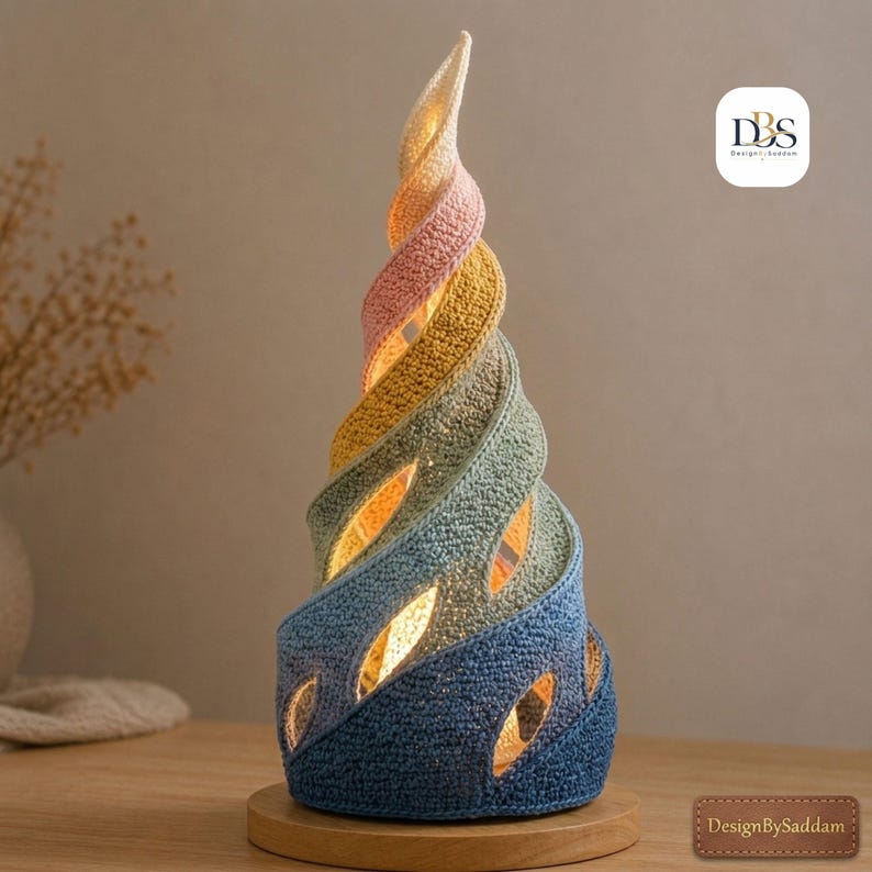 May include: A spiral-shaped table lamp featuring a textured, crocheted exterior in a gradient of blue, green, yellow, and pink. The structure has open gaps that emit a warm internal light, resting on a circular wooden base with the text DesignBySaddam.