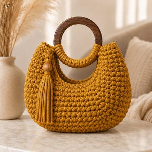 Peut inclure: Un sac à main en crochet jaune moutarde doté d'une anse circulaire en bois brun foncé et d'un pompon assorti attaché sur le côté. Le sac présente un motif de mailles nouées texturées et une forme arrondie en demi-lune.