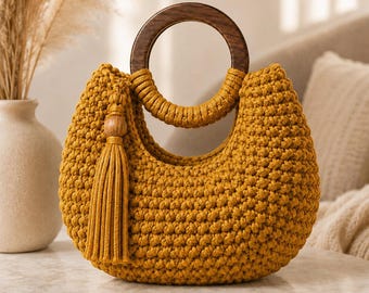 Puede incluir: Un bolso de ganchillo color amarillo mostaza con un asa circular de madera marrón oscuro y una borla a juego unida al lateral. El bolso presenta un patrón de punto anudado texturizado y una forma redondeada de media luna.