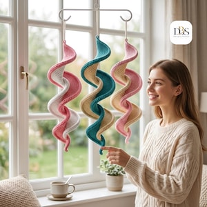 Könnte beinhalten: Drei hängende Spiraldekorationen aus Strickstoff, die an einem weißen Metallrahmen befestigt sind. Die Spiralen zeigen Farbkombinationen aus Rosa und Weiß, Petrol und Beige sowie Rosa und Beige. Der Text DBS Design-Garden ist sichtbar.