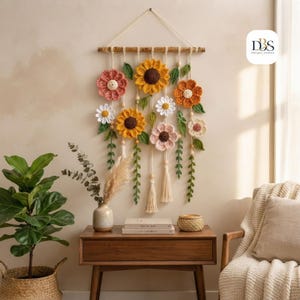 May include: A wall hanging featuring crocheted flowers in shades of yellow, orange, pink, and white, accented with green crocheted leaves and vines. The flowers are suspended from a wooden dowel by cream-colored cords with wooden beads and tassels.