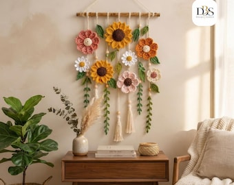 Puede incluir: Un tapiz de pared con flores de ganchillo en tonos amarillo, naranja, rosa y blanco, acentuado con hojas y enredaderas de ganchillo verdes. Las flores cuelgan de un palo de madera mediante cordones color crema con cuentas de madera y borlas.