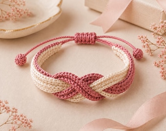 Puede incluir: Una pulsera de cordón trenzado con un diseño de nudo infinito en colores alternos rosa empolvado y crema. La pulsera incluye un cierre deslizante ajustable con dos pequeños extremos esféricos, mostrando una construcción trenzada texturizada.