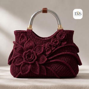 May include: A deep burgundy crochet handbag featuring a large dimensional floral applique on the front, a curved top opening, and a silver-toned metal handle with a brown leather accent. The logo DBS Design Blossom is visible in the top right corner.