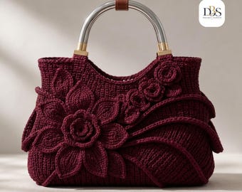 Puede incluir: Un bolso de ganchillo color burdeos intenso con una gran aplicación floral tridimensional en la parte delantera, una abertura superior curva y un asa de metal plateado con un detalle de cuero marrón. El logo DBS Design Blossom es visible.