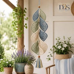 May include: A hanging crochet mobile featuring a vertical arrangement of six leaf-shaped elements in shades of sage green, dusty blue, and cream. The leaves are connected by a central cord with wooden beads and finished with a multi-colored tassel.