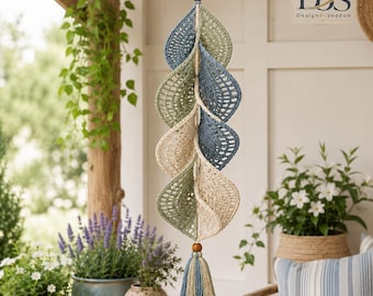 May include: A hanging crochet mobile featuring a vertical arrangement of six leaf-shaped elements in shades of sage green, dusty blue, and cream. The leaves are connected by a central cord with wooden beads and finished with a multi-colored tassel.