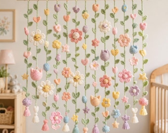 Puede incluir: Un tapiz de pared con múltiples hilos verticales de flores, hojas, corazones y estrellas de ganchillo en colores pastel, suspendidos de una varilla de madera. Cada hilo termina en una pequeña borla y varias flores tienen caras bordadas.