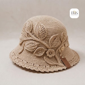 Peut inclure: Un chapeau bob en crochet beige orné d'un motif appliqué de feuilles et de fleurs texturées. Le chapeau présente un bord festonné et une petite languette en similicuir marron. Le logo DBS Design-Sodden est visible dans le coin supérieur droit.