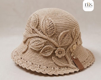 Puede incluir: Un sombrero de pescador de ganchillo beige con un diseño de apliques de hojas y flores texturizadas. El sombrero tiene un borde festoneado y un pequeño detalle de cuero marrón. El logotipo DBS Design-Sodden es visible en la esquina superior derecha.