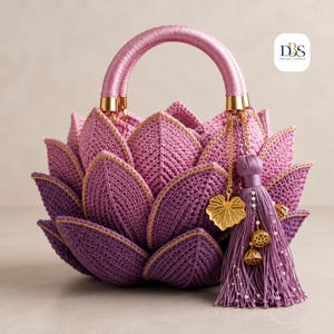 May include: A crochet handbag shaped like a lotus flower in shades of purple and pink. The bag features layered petals, a wrapped handle, and a decorative tassel with gold-toned leaf and lotus pod charms. The logo DBS Design-Season is visible.