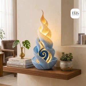 Könnte beinhalten: Eine spiralförmige Tischleuchte mit strukturierter Oberfläche, die von Weiß an der Spitze zu Hellblau und Dunkelblau an der Basis verläuft. Die Lampe strahlt ein warmes inneres Licht aus und zeigt das Logo DBS Design-Goddess oben rechts.