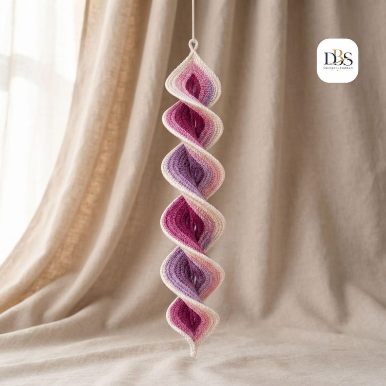 May include: A hanging spiral ornament crafted from crochet yarn, featuring a gradient color scheme that transitions from cream to light pink, magenta, and deep purple. The structure is twisted into a vertical helix shape with a loop at the top.